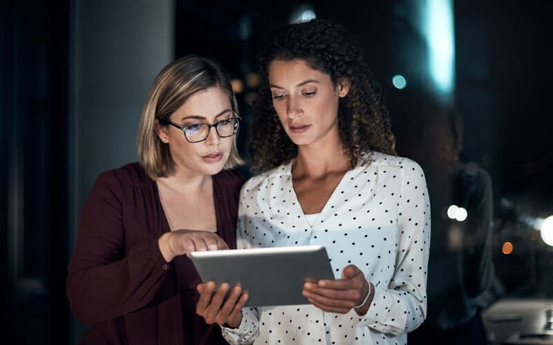 Two women using tablet device late at night in office